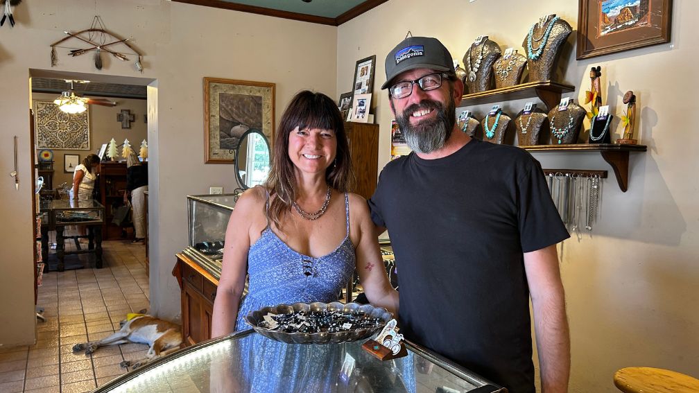 Two people stand behind a glass jewelry display case in a Mesilla shop, smiling at the camera. Authentic silver necklaces and jewelry adorn the walls and shelves. A dog lies on the floor in the background.